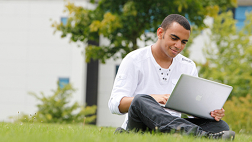 a students sits on the grass with his laptop, making notes 