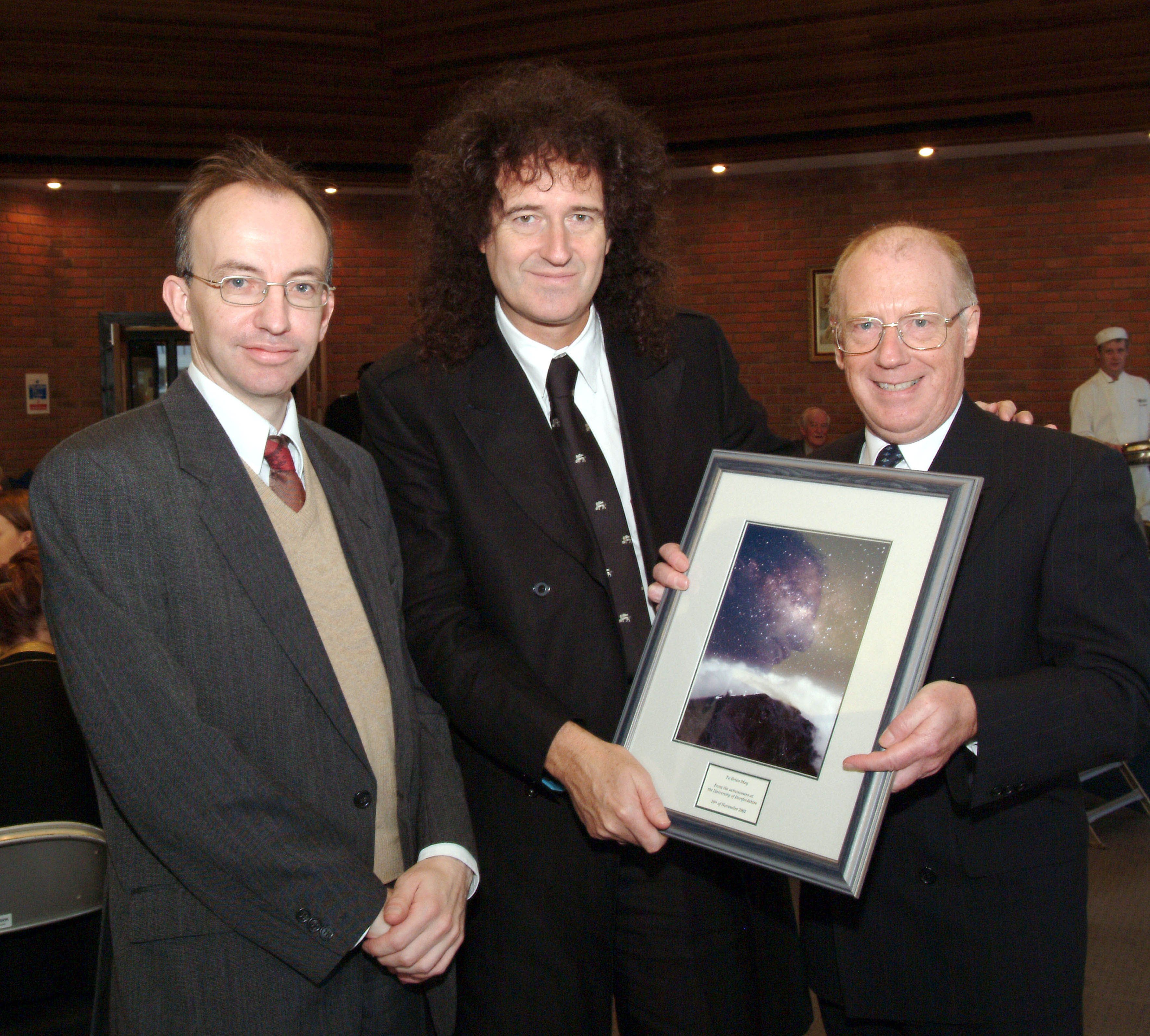 Brian May The photo shows Brian May being presented with an astronomical photograph by James Hough and colleague James Collett after the honorary degree ceremony.
